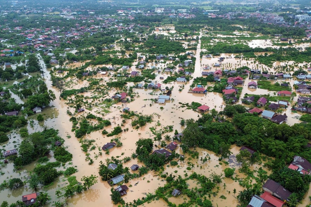 Foto udara yang memperlihatkan pemandangan luas banjir bandang yang menenggelamkan kawasan permukiman di pedesaan Sumatera. Air banjir berwarna cokelat keruh merendam jalanan, bangunan, dan area sekitarnya, dengan latar belakang perbukitan yang tertutup awan mendung