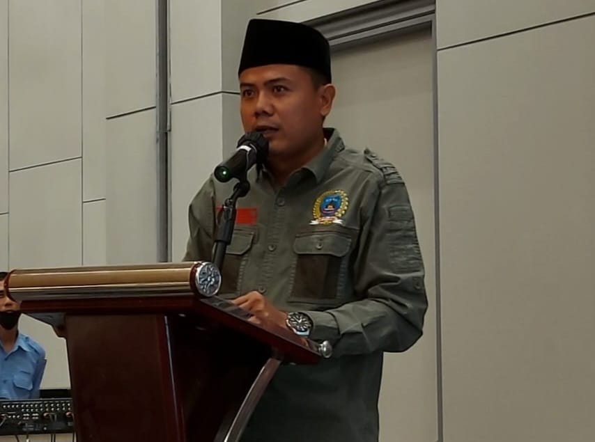 Ketua Umum Aliansi Madura Indonesia (AMI), Baihaki Akbar, mengenakan peci hitam dan seragam organisasi berwarna hijau zaitun, sedang berbicara di podium menggunakan mikrofon dalam sebuah acara resmi.