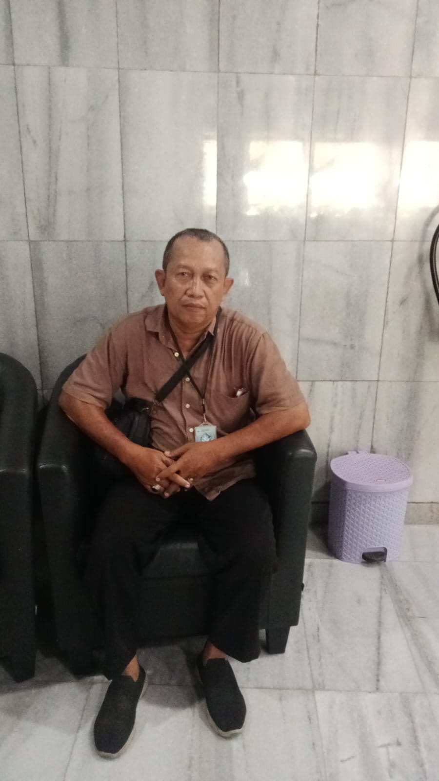 Ir. Bagus Prihatin, Komisaris PT Tabir Lentera Nusantara, mengenakan kemeja cokelat dan duduk di kursi hitam, menatap lurus ke depan dengan ekspresi serius di depan dinding marmer abu-abu.
