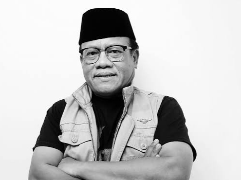 "Potret formal seorang pria mengenakan peci hitam dan rompi dengan lencana sayap, berdiri dengan tangan terlipat di dada, latar belakang putih"