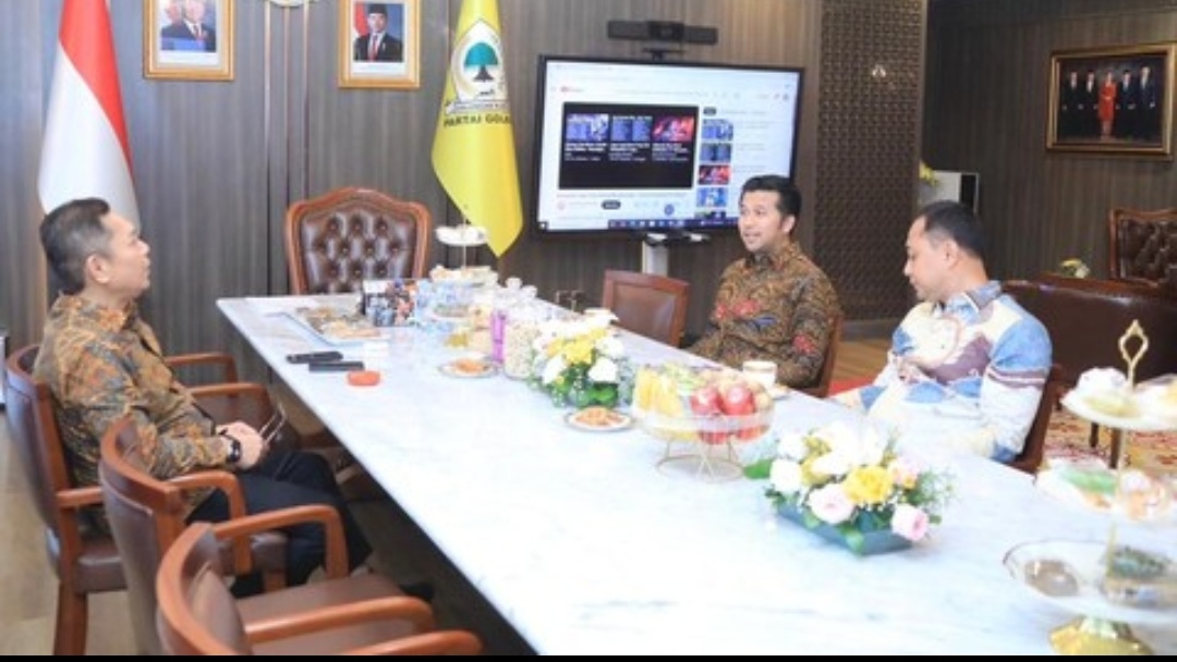 Tiga pejabat pria berbatik sedang duduk mengelilingi meja marmer panjang yang dipenuhi makanan dan minuman ringan dalam sebuah ruangan kantor formal. Di sisi kiri meja, seorang pria paruh baya mengenakan batik cokelat gelap sedang berbicara sambil bersandar. Di seberangnya, di tengah meja, seorang pria muda mengenakan batik cokelat bermotif tersenyum dan mendengarkan. Di ujung kanan meja, pria ketiga mengenakan batik biru cerah juga terlihat fokus. Di latar belakang terdapat bendera Indonesia, bendera Partai Golkar, dan layar TV besar menampilkan video."