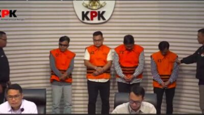 Tersangka kasus korupsi proyek PUPR OKU dihadirkan KPK dalam konferensi pers. Empat pria mengenakan rompi tahanan oranye berangka berdiri di podium, diapit oleh petugas KPK. Terdapat logo KPK besar di latar belakang