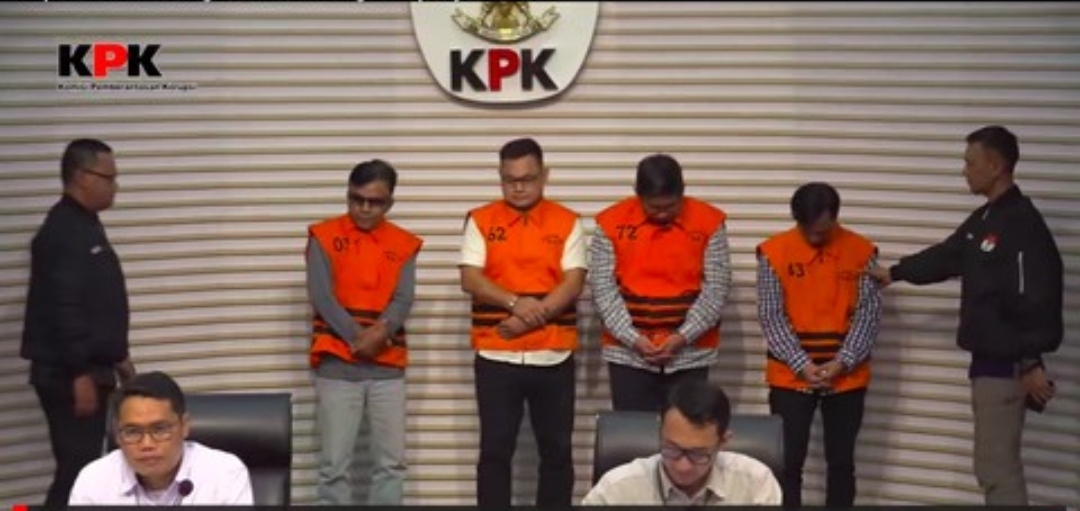 Tersangka kasus korupsi proyek PUPR OKU dihadirkan KPK dalam konferensi pers. Empat pria mengenakan rompi tahanan oranye berangka berdiri di podium, diapit oleh petugas KPK. Terdapat logo KPK besar di latar belakang