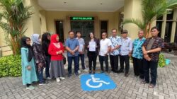 Sekelompok warga, termasuk Wakil Wali Kota Surabaya Armuji dan seorang pengacara wanita, berpose di depan gedung SPKT Polda Jatim. Mereka memegang dokumen laporan polisi dan berdiri di samping simbol kursi roda di area parkir, menandakan pelaporan kasus penipuan properti cessie.