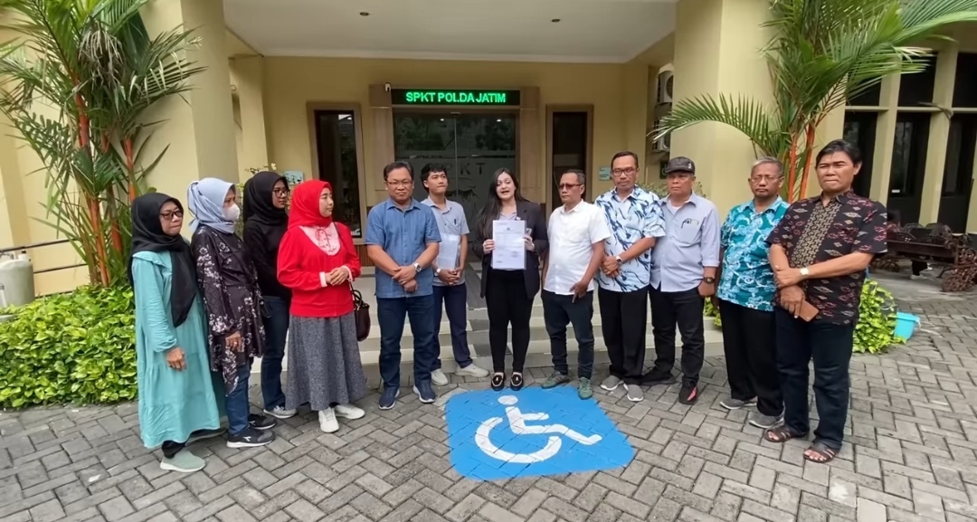 Sekelompok warga, termasuk Wakil Wali Kota Surabaya Armuji dan seorang pengacara wanita, berpose di depan gedung SPKT Polda Jatim. Mereka memegang dokumen laporan polisi dan berdiri di samping simbol kursi roda di area parkir, menandakan pelaporan kasus penipuan properti cessie.