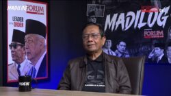 Seorang pria paruh baya mengenakan jaket kulit cokelat dan kaus hitam duduk di kursi kulit gelap di sebuah studio podcast. Di belakangnya terdapat dua poster besar dengan tulisan "MADILOG" dan "FORUM KEADILAN". Pria tersebut tersenyum tipis ke arah kamera, di meja depannya ada cangkir hitam bertuliskan "MADILOG".