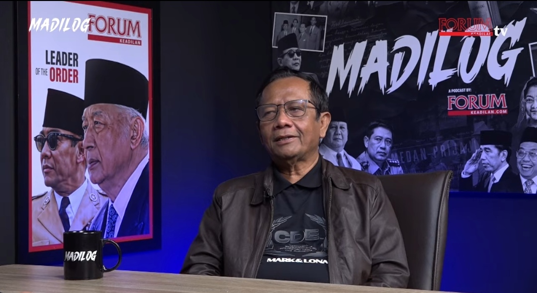 Seorang pria paruh baya mengenakan jaket kulit cokelat dan kaus hitam duduk di kursi kulit gelap di sebuah studio podcast. Di belakangnya terdapat dua poster besar dengan tulisan "MADILOG" dan "FORUM KEADILAN". Pria tersebut tersenyum tipis ke arah kamera, di meja depannya ada cangkir hitam bertuliskan "MADILOG".