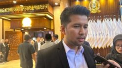 Wakil Gubernur Jawa Timur, Emil Elestianto Dardak, mengenakan jas hitam dan kemeja putih, sedang memberikan keterangan pers kepada wartawan dengan latar belakang interior gedung DPRD Jawa Timur yang bernuansa kayu dan lambang emas.