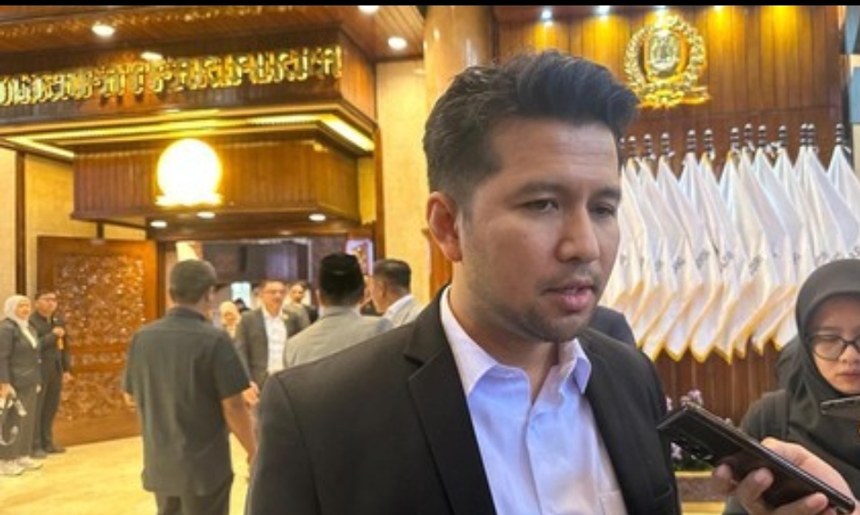 Wakil Gubernur Jawa Timur, Emil Elestianto Dardak, mengenakan jas hitam dan kemeja putih, sedang memberikan keterangan pers kepada wartawan dengan latar belakang interior gedung DPRD Jawa Timur yang bernuansa kayu dan lambang emas.