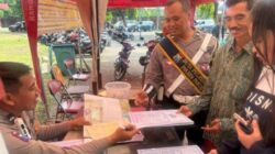 Seorang petugas polisi berseragam lengkap dengan selempang bertuliskan "Duta Pelayanan Polres Pasuruan" sedang mendampingi dan membantu dua orang warga dalam proses verifikasi dokumen di meja layanan luar ruangan Kantor Bersama Samsat Kota Pasuruan.