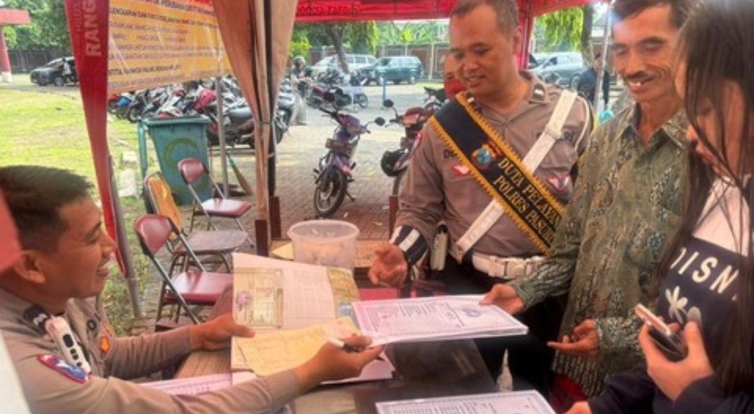 Seorang petugas polisi berseragam lengkap dengan selempang bertuliskan "Duta Pelayanan Polres Pasuruan" sedang mendampingi dan membantu dua orang warga dalam proses verifikasi dokumen di meja layanan luar ruangan Kantor Bersama Samsat Kota Pasuruan.