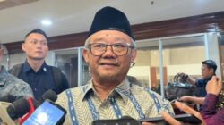 Mendikdasmen Abdul Mu'ti mengenakan kemeja batik dan peci hitam, sedang berbicara di podium dengan latar belakang spanduk Musyawarah Nasional XX IKAPI Tahun 2025.