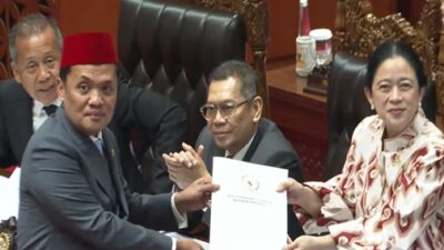 Puan Maharani, Ketua DPR RI, mengenakan blus batik cokelat dan merah, menerima berkas dokumen dari Ketua Komisi III DPR, Habiburrahman, yang mengenakan setelan jas dan peci merah. Mereka berdiri di meja pimpinan dalam rapat paripurna. Di belakang mereka, anggota DPR lain terlihat ikut bertepuk tangan. Logo "TNP Parlemen LANGSUNG" dan jendela penerjemah bahasa isyarat terlihat di sudut kanan atas dan bawah.