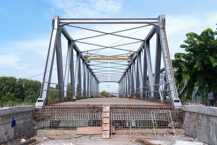 Perspektif lorong jembatan rangka baja yang lantainya dipenuhi anyaman besi beton (rebar) siap cor, dengan pemandangan langit biru dan pepohonan di sisi kanan, menunjukkan tahap persiapan pengecoran lantai jembatan.