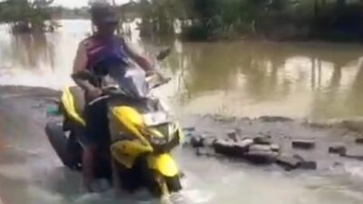 Seorang pria mengendarai sepeda motor kuning menerobos jalan desa yang tergenang air banjir dengan latar belakang sawah dan pepohonan.