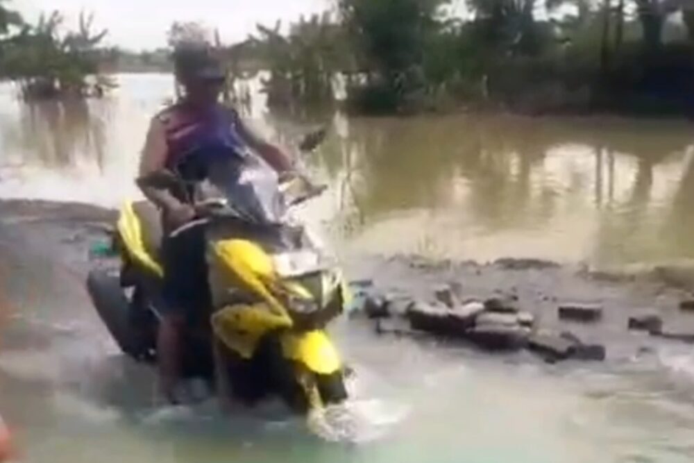 Seorang pria mengendarai sepeda motor kuning menerobos jalan desa yang tergenang air banjir dengan latar belakang sawah dan pepohonan.