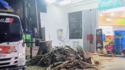 Suasana malam hari di depan toko Alfa Midi yang terang benderang. Terlihat tumpukan besar potongan batang kayu dan sisa penebangan pohon di area parkir yang berantakan. Sebagian badan truk pengangkut berwarna putih terlihat di sisi kiri gambar."