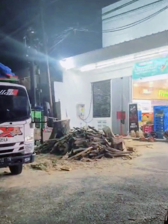Suasana malam hari di depan toko Alfa Midi yang terang benderang. Terlihat tumpukan besar potongan batang kayu dan sisa penebangan pohon di area parkir yang berantakan. Sebagian badan truk pengangkut berwarna putih terlihat di sisi kiri gambar."