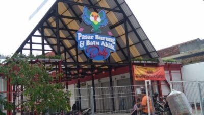Foto tampak depan gerbang bangunan Pasar Burung dan Batu Akik Dolly di Surabaya dengan atap rangka baja yang tinggi. Terlihat papan nama besar dengan ikon burung, pagar besi pembatas, dan spanduk oranye bertuliskan 'Posko Penempatan Calon Pedagang'. Seorang warga tampak melintas di depan pagar menggunakan sepeda motor membawa muatan barang.