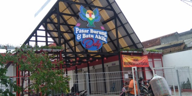 Foto tampak depan gerbang bangunan Pasar Burung dan Batu Akik Dolly di Surabaya dengan atap rangka baja yang tinggi. Terlihat papan nama besar dengan ikon burung, pagar besi pembatas, dan spanduk oranye bertuliskan 'Posko Penempatan Calon Pedagang'. Seorang warga tampak melintas di depan pagar menggunakan sepeda motor membawa muatan barang.