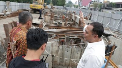Wali Kota Surabaya, Eri Cahyadi (berkemeja putih di kanan), memberikan instruksi sambil menunjuk ke dalam galian proyek pembangunan rumah pompa yang dalam dan kompleks. Ia didampingi dua orang di tepi area konstruksi yang dipenuhi struktur beton bertulang, balok baja penyangga, dan sebuah alat berat ekskavator di latar belakang.