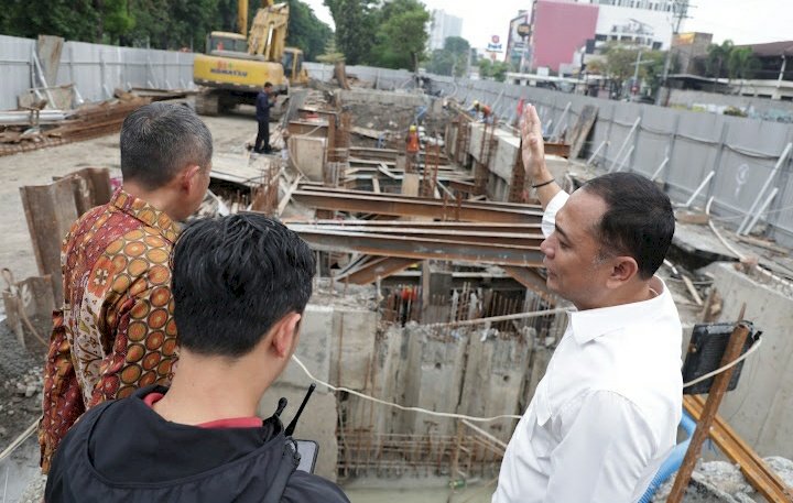 Wali Kota Surabaya, Eri Cahyadi (berkemeja putih di kanan), memberikan instruksi sambil menunjuk ke dalam galian proyek pembangunan rumah pompa yang dalam dan kompleks. Ia didampingi dua orang di tepi area konstruksi yang dipenuhi struktur beton bertulang, balok baja penyangga, dan sebuah alat berat ekskavator di latar belakang.