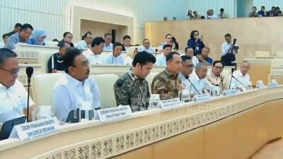 Sebuah ruangan rapat besar dengan deretan meja panjang, tempat sejumlah pejabat dan peserta RDP sedang duduk. Mereka mengenakan kemeja dan batik, fokus pada pembahasan di depan. Tampak layar proyektor kecil di setiap meja dan latar belakang dinding berwarna krem dengan aksen kayu.