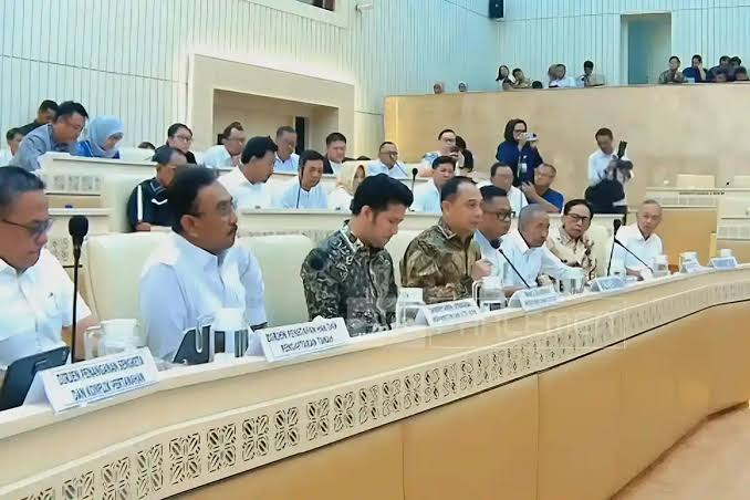 Sebuah ruangan rapat besar dengan deretan meja panjang, tempat sejumlah pejabat dan peserta RDP sedang duduk. Mereka mengenakan kemeja dan batik, fokus pada pembahasan di depan. Tampak layar proyektor kecil di setiap meja dan latar belakang dinding berwarna krem dengan aksen kayu.