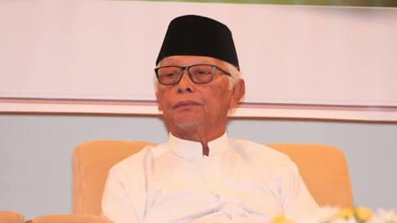Foto Wakil Rais Aam PBNU, KH Anwar Iskandar, sedang duduk santai di sofa krem. Ia mengenakan baju koko putih, peci hitam, dan kacamata, dengan ekspresi tenang namun serius.