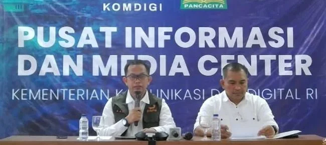 Dua pria duduk di meja konferensi pers berlatar spanduk biru bertuliskan Pusat Informasi dan Media Center Kementerian Komunikasi dan Digital RI dengan logo Komdigi dan Pancacita. Pria berkacamata yang mengenakan rompi BNPB di sebelah kiri tampak berbicara menggunakan mikrofon, sementara pria berbaju putih di sebelahnya sedang membaca dokumen.