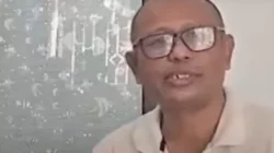 Seorang pria berkacamata dengan kepala plontos dan mengenakan kemeja polo berwarna krem terlihat berbicara menghadap kamera.