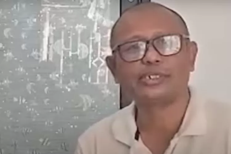 Seorang pria berkacamata dengan kepala plontos dan mengenakan kemeja polo berwarna krem terlihat berbicara menghadap kamera.