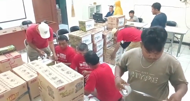 Sejumlah pria, sebagian mengenakan kaos merah bertuliskan Rukun Warga 03, sedang sibuk mengemas tumpukan kardus bantuan logistik menggunakan lakban di dalam ruangan Balai RW