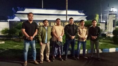 Enam orang pria berdiri berjejer menghadap kamera di depan gerbang masuk bertuliskan Rumah Dinas Bupati Sampang pada malam hari.