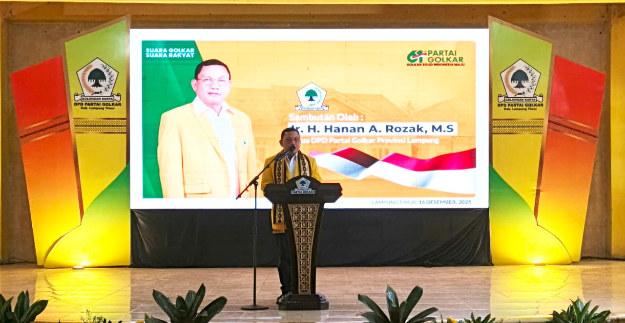 Seorang pria berdiri di podium menyampaikan sambutan pada acara Musda IV Partai Golkar Lampung Timur, dengan latar layar besar menampilkan nama H. Hanan A. Rozak dan atribut Partai Golkar.