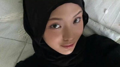 Foto close-up seorang wanita muda mengenakan hijab hitam dan pakaian berwarna senada sedang melakukan swafoto dengan ekspresi wajah tenang menatap kamera. Latar belakang foto memperlihatkan permukaan kain berwarna abu-abu muda dengan tekstur lembut.