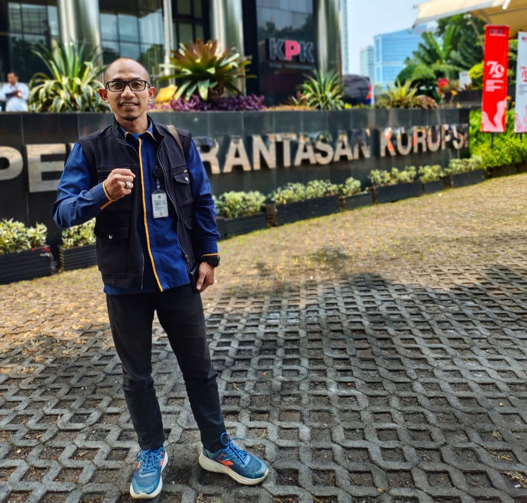 Seorang pria berkacamata dengan rompi hitam dan kemeja biru berdiri sambil mengepalkan tangan di depan gedung Komisi Pemberantasan Korupsi (KPK).