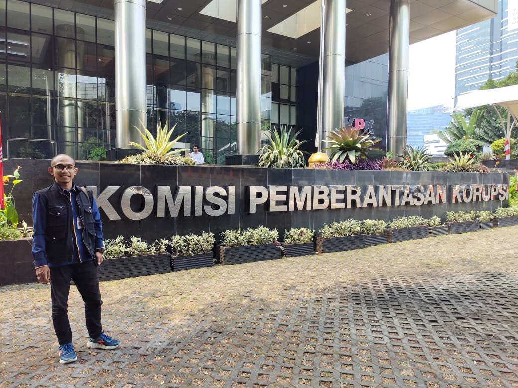 Seno Aji, S.Sos., S.H., M.H., berdiri di depan papan nama bertuliskan Komisi Pemberantasan Korupsi (KPK) dengan latar gedung tinggi.
