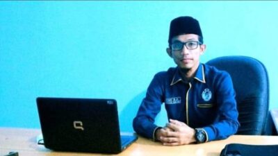 Seorang pria mengenakan peci dan kemeja biru duduk di meja kerja dengan laptop di depannya.