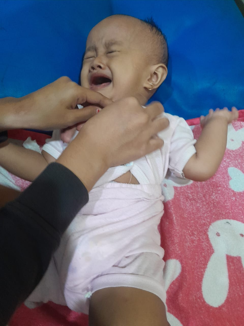 Seorang bayi tampak menangis saat berada di atas alas, dengan tangan orang dewasa terlihat memegang bagian tubuh bayi tersebut.