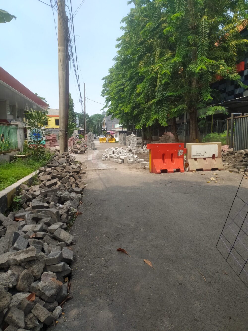 Foto sudut pandang luas (wide shot) yang memperlihatkan kondisi jalan lingkungan yang sedang dalam masa konstruksi namun terlihat sepi. Di sisi kiri depan terdapat tumpukan besar puing-puing paving atau aspal lama yang berwarna gelap. Di tengah jalan berdiri pembatas jalan (road barrier) plastik berwarna oranye dan putih. Jauh di latar belakang jalan, terlihat tumpukan paving block baru berwarna abu-abu terang. Tidak ada aktivitas pekerja atau papan nama proyek yang terlihat di sepanjang jalan yang dinaungi pohon-pohon besar di sisi kanan ini.