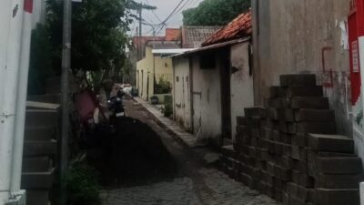 Kondisi gang sempit di Jalan Tanjungsari V, Surabaya, yang sedang dalam pengerjaan proyek drainase. Terlihat tumpukan material beton saluran di kedua sisi jalan dan gundukan tanah galian di tengah jalan yang menghambat akses.