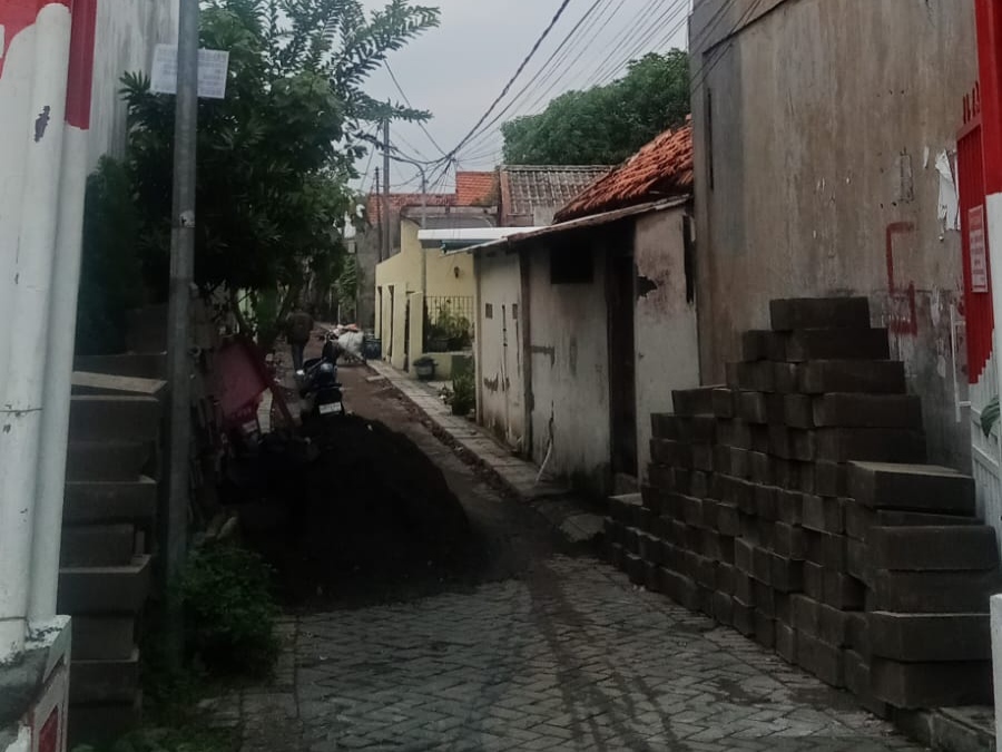 Kondisi gang sempit di Jalan Tanjungsari V, Surabaya, yang sedang dalam pengerjaan proyek drainase. Terlihat tumpukan material beton saluran di kedua sisi jalan dan gundukan tanah galian di tengah jalan yang menghambat akses.