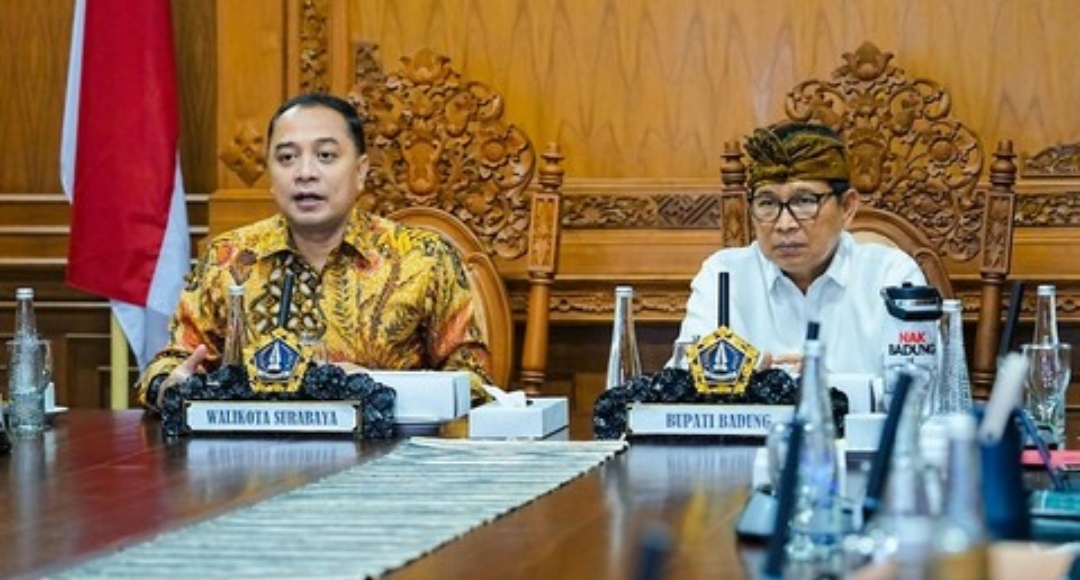 Wali Kota Surabaya Eri Cahyadi mengenakan batik dan Bupati Badung I Wayan Adi Arnawa mengenakan udeng duduk berdampingan di meja rapat dengan papan nama jabatan terlihat jelas di depan mereka.