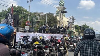 Suasana aksi unjuk rasa anti-korupsi di jalan raya, terlihat panggung dengan spanduk bertuliskan 'TOLAK KORUPSI' dan kerumunan sepeda motor yang menyebabkan kemacetan.