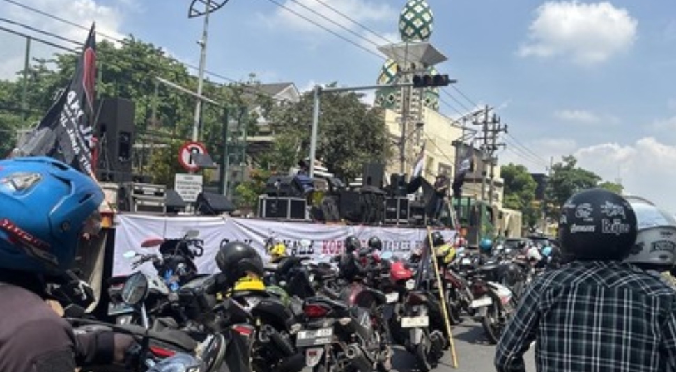 Suasana aksi unjuk rasa anti-korupsi di jalan raya, terlihat panggung dengan spanduk bertuliskan 'TOLAK KORUPSI' dan kerumunan sepeda motor yang menyebabkan kemacetan.