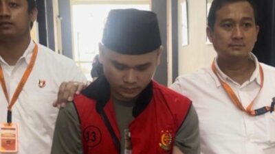 Tersangka Alvi Maulana mengenakan rompi tahanan merah dan peci hitam dikawal oleh dua petugas kejaksaan.