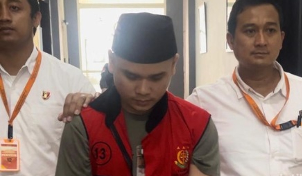 Tersangka Alvi Maulana mengenakan rompi tahanan merah dan peci hitam dikawal oleh dua petugas kejaksaan.