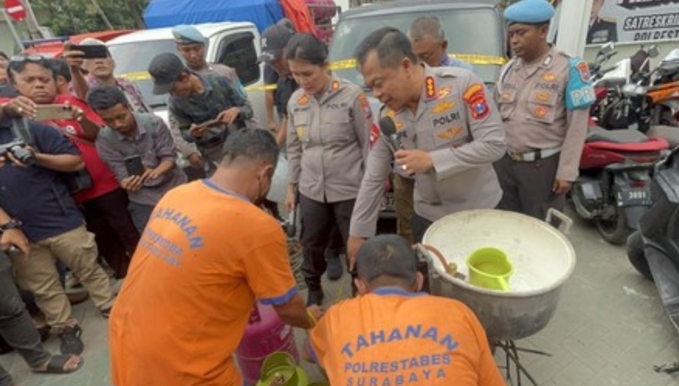 Dua tersangka berbaju tahanan oranye memperagakan pengoplosan gas elpiji di hadapan polisi dan wartawan saat konferensi pers. Barang bukti berupa tabung gas 3 kg, tabung 12 kg, dan panci besar terlihat digelar di lokasi kejadian.