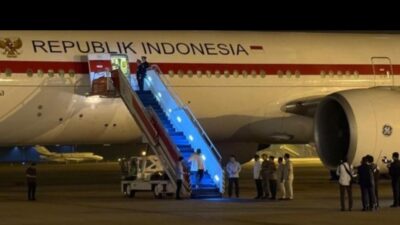 Pesawat kepresidenan bertuliskan Republik Indonesia parkir di landasan pacu pada malam hari saat seseorang menuruni tangga pesawat yang diterangi lampu biru. Di bawah tangga, sekelompok pejabat dan petugas berdiri berbaris menyambut kedatangan tersebut.